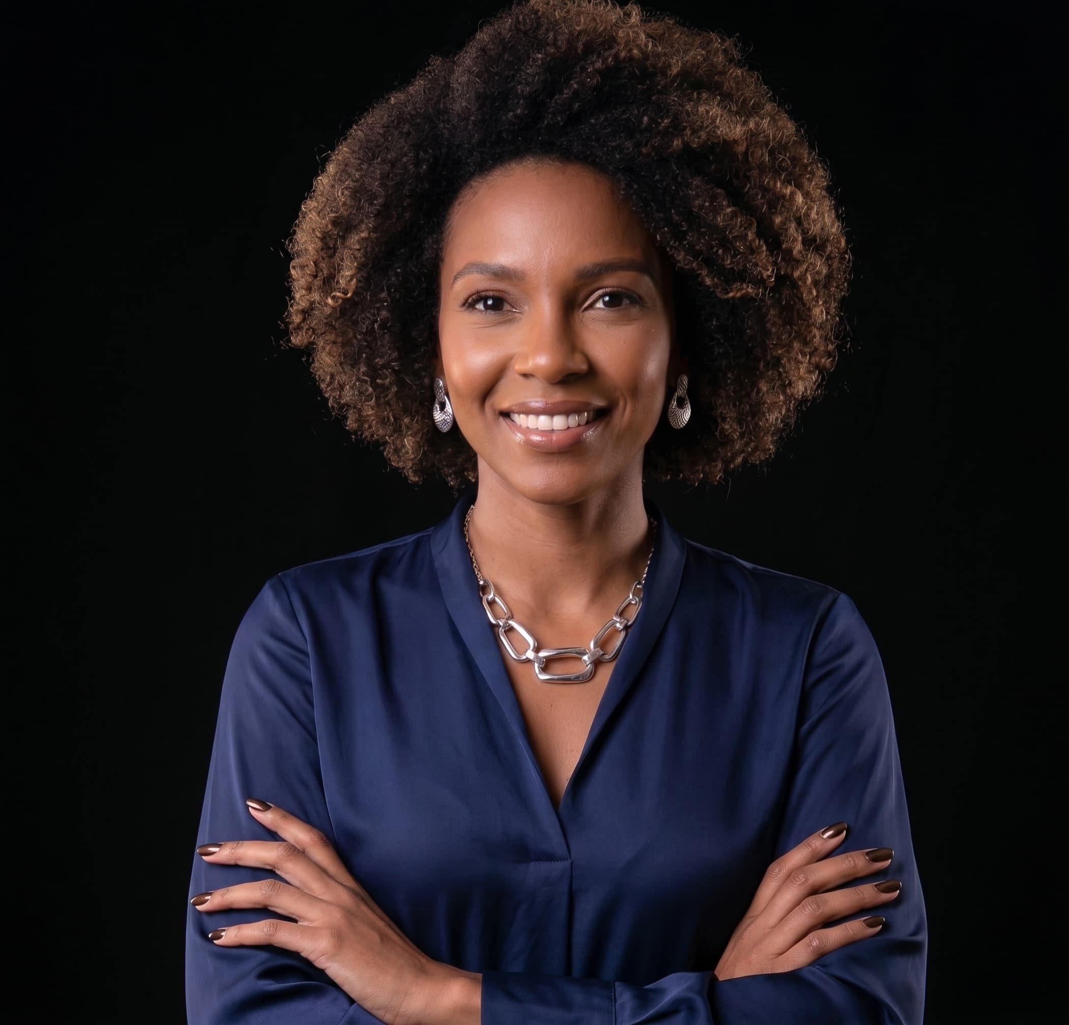 Beatriz Nogueira em retrato de estúdio sobre fundo preto, de blusa azul-marinho, cabelo cacheado volumoso e braços cruzados.
