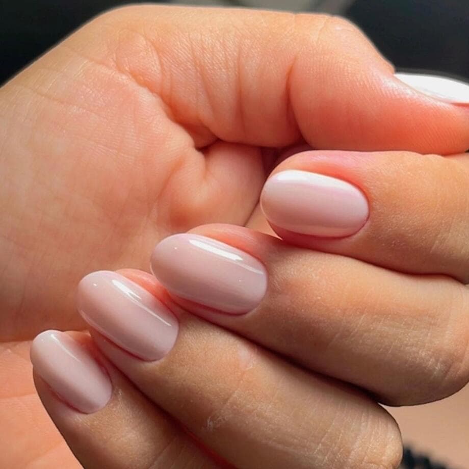 Close de uma mão com unhas ovais curtas, esmaltadas em gel rosa nude com acabamento brilhante.