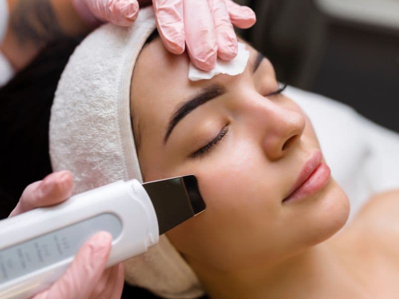 Cliente deitada, de olhos fechados e faixa branca no cabelo, recebendo limpeza de pele com espátula ultrassônica na bochecha.